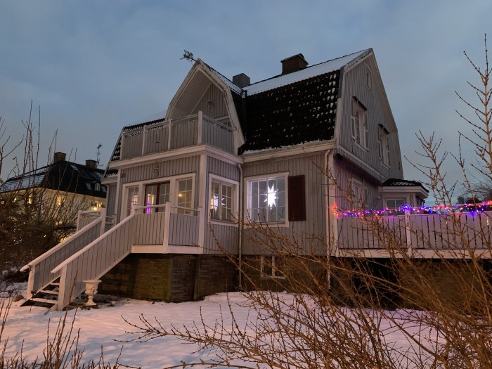 Hus med sekelskiftesstil i skymningen med julbelysning och snö på marken.