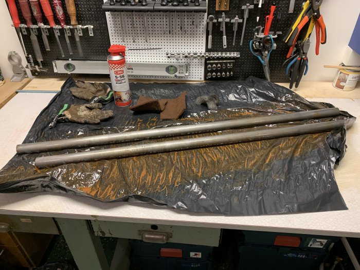 Rostiga rör på plastöverdrag med en flaska rostlösare och handskar på verkstadsbänk.