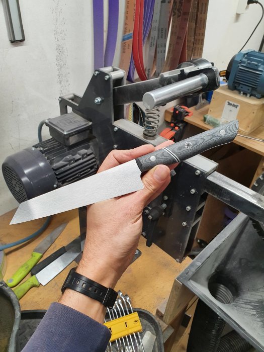 Hand som håller en stort kökskniv framför en slipmaskin i en verkstad.