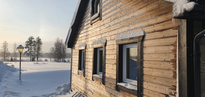 Vinterbild av trähus med snö på taket och fönsterbrädorna, gatlykta tänt i förgrunden.