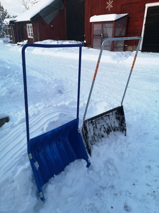 Två snöskyfflar står i snön, en ny blå Fiskars och en äldre sliten svart från Biltema.