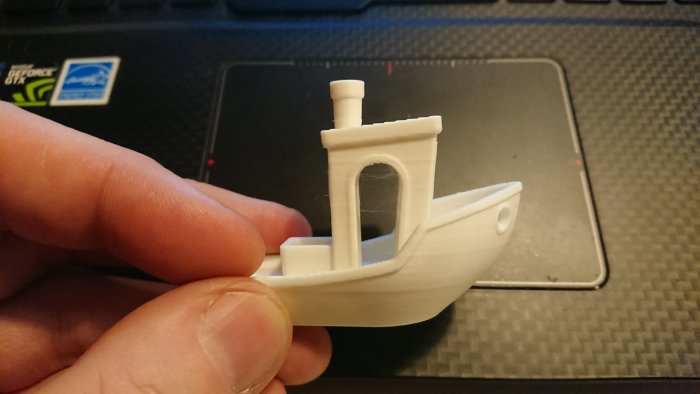 3D-utskriven vit båtmodell (Benchy) som hålls mellan fingrarna mot en bärbar dators tangentbord.