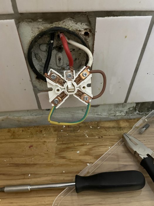 Öppen eluttag med felkopplade trådar och kakel, verktyg syns på golvet.