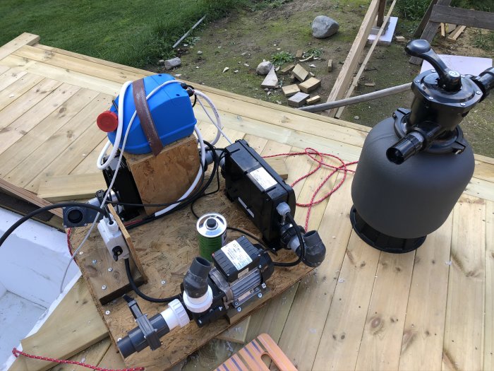 Reningsutrustning för terasspool med pump, filter och slangar monterade på träplattform.