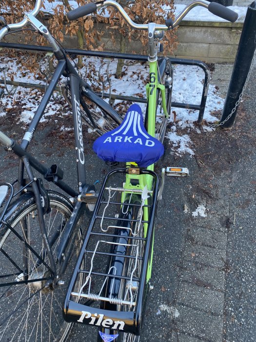 Grön cykel med märket Pilen och en blå sadelskydd där det står ARKAD, parkerad bredvid en annan cykel.