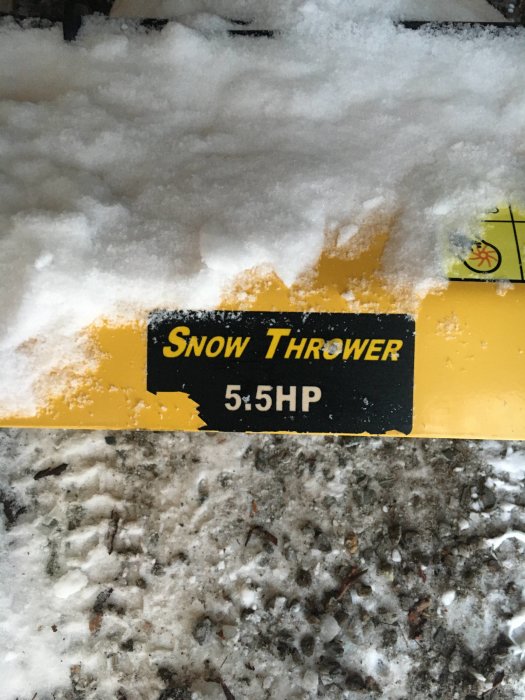 Delvis täckt skylt på en snöslunga märkt "Snow Thrower 5.5HP" omgiven av snö och smutsig is.