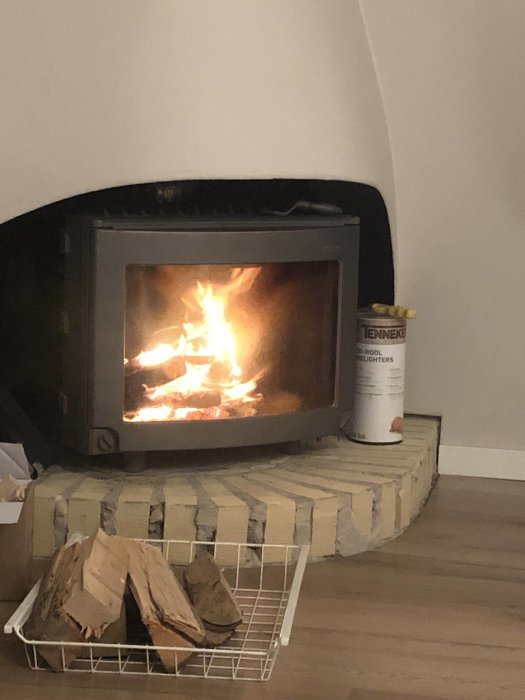 Eldstad med brinnande ved i ett hem, vedkorg och tändsticksburk bredvid, i ett försök att sänka elräkningen.