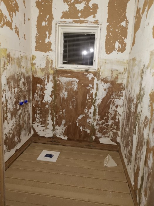 Ett orenoverat badrum med exponerat golvspån, väggar i behov av renovering och ett fönster.