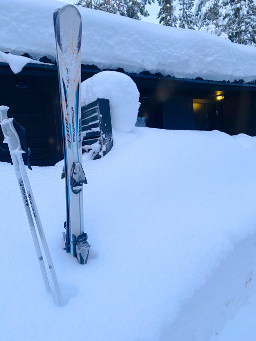 Alpina skidor upprätt i djup snö framför snötäckt hus med belysning i Lindvallen.