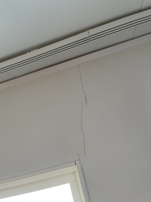 Sprickor i hörnet där en vit vägg och tak möts, nära lister och en fönsterkarm.