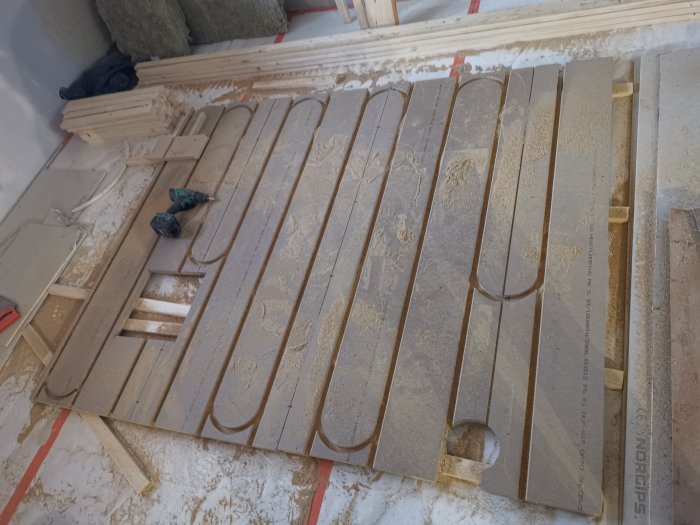 Nyfrästa spår i trägolv för installation av golvvärme med synliga isoleringsmaterial och mäthjälpmedel.
