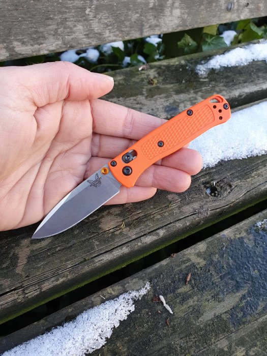 Hand håller en uppfälld fällkniv med orange handtag på en snötäckt träbänk.