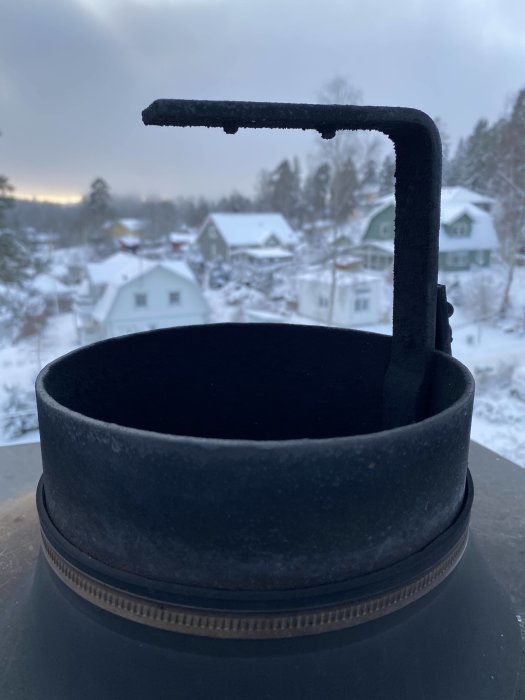 Rund skorstensskyddsplåt med fästanordning framför snöklädda hus.