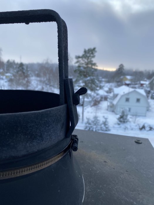 Närbild av en skorsten med lossnat skydd och enkel rund plåt mot en snöig landskapsbakgrund.