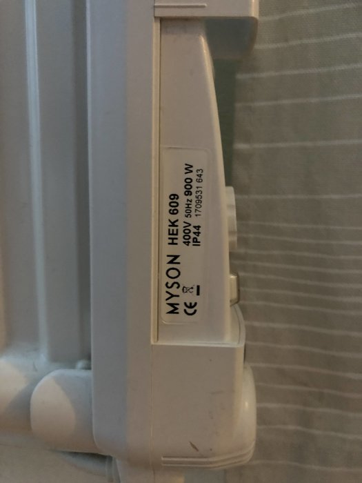Myson MEK 609 elradiator med identifieringsetikett och termostatknappar, delvis skymd av gardin.