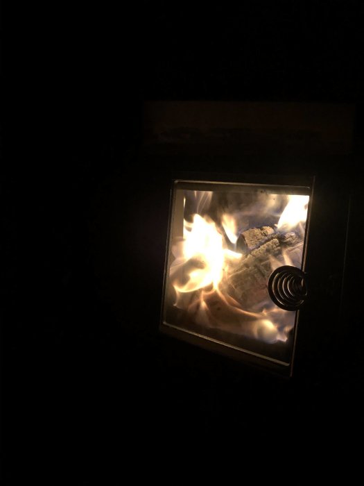 Brinnande ved i en bastuugn sedd genom en glaslucka i ett mörkt rum.