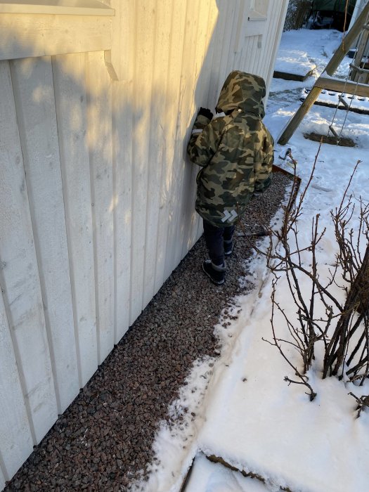 Person i kamouflagejacka drar kabel längs med ett vitt hus, snö på marken.