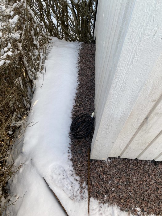 Kabel upprullad vid hörnet av ett hus med snötäckt mark och buskar.