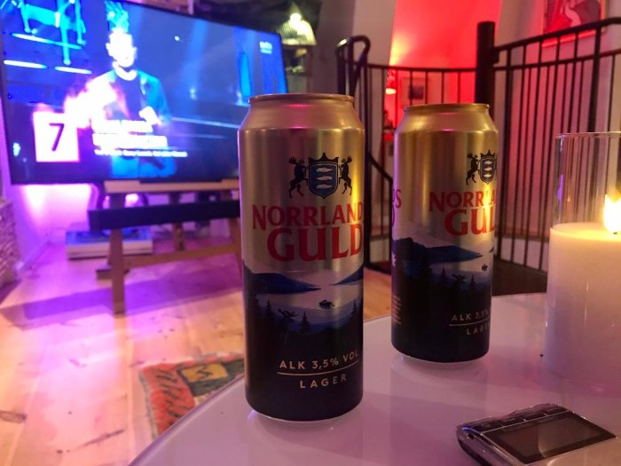 Två burkar Norrlands Guld lager framför en tänd ljusstake och suddig TV-skärm i bakgrunden.