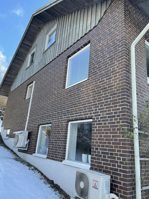 Husfasad med tegel och träbeklädnad, fönster och värmepumpar, snötäckt mark och synligt stuprör.