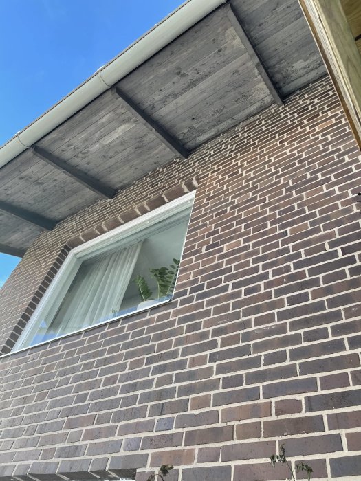 Del av husfasad med tegelsten och fönster under träbjälklag med plats för svarta stuprör.