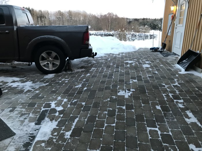 Stenlagd uppfart med delvis bortblåst snö, pickup-truck till vänster och snöskyfflar mot husväggen.