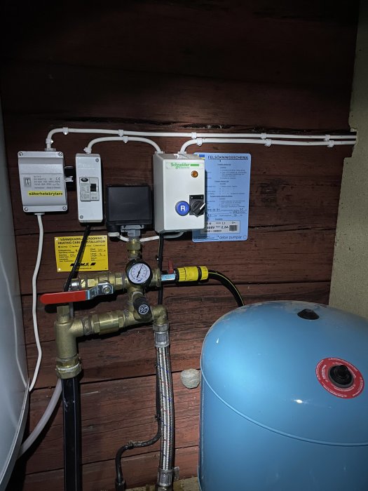 Hydrofor och säkerhetsutrustning för vattensystem i en stuga, med kopplingar, rör och en tryckmätare.