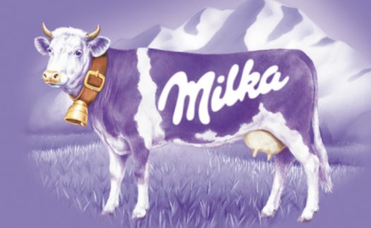 Milka-ko med lila och vit päls står framför berg, med ett stort "Milka" på sin kropp och en klocka runt halsen.