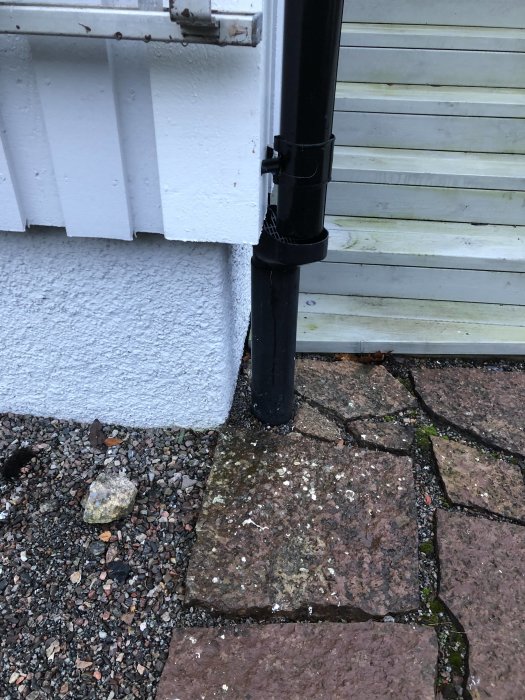 Hörnet på ett hus med vit putsad fasad och ett stuprör nära marksten och grus.