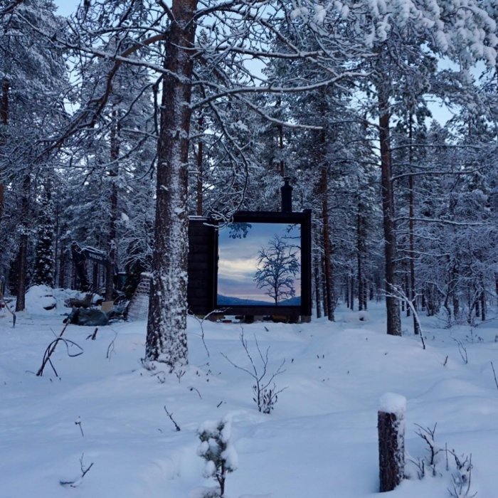 Litet svart förkolnat bastuhus i snötäckt skog, med vedeldad skorsten och inspiration från finska bastustugor.