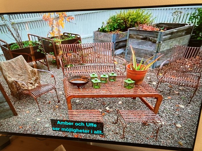 Rustik utemöbelgrupp med bord, stolar och bänk i smide på grusunderlag, visad på TV-skärm.