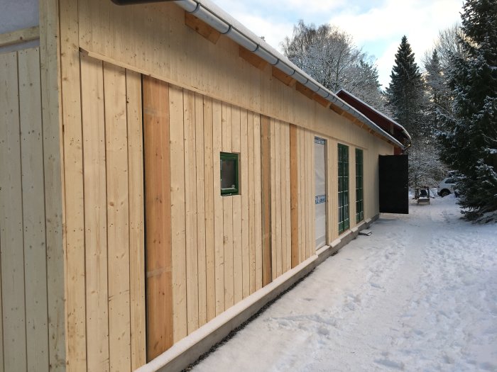 Träfasad på byggnad med varierande bredder på panelerna, snötäckt mark och träd i bakgrunden.