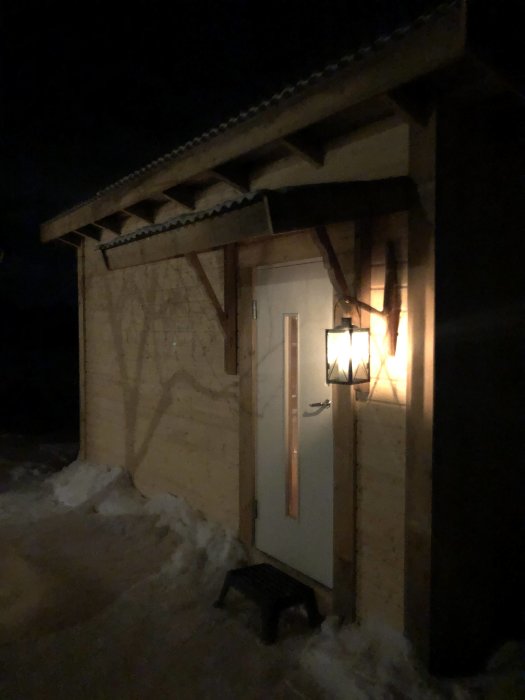 Bastu i vintermiljö nattetid med tänd lampa vid dörren och snö runtomkring.