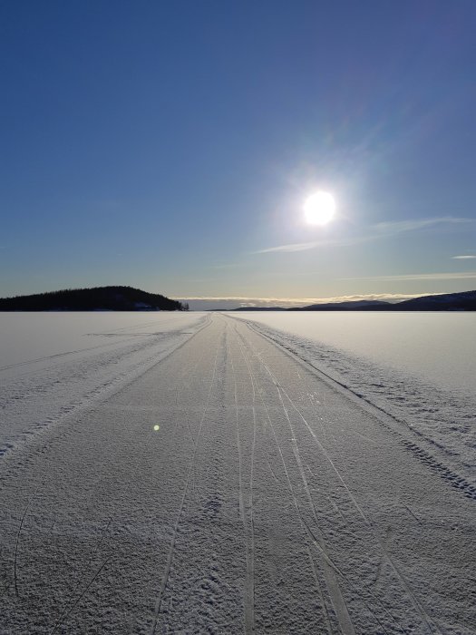 Solig vinterscen med skidspår på en frusen sjö omgiven av snöklädda landskap och klarblå himmel.