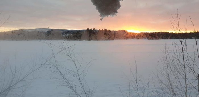Solnedgång sett genom ett fönster med fukt, med snötäckt landskap och träd i silhuett.