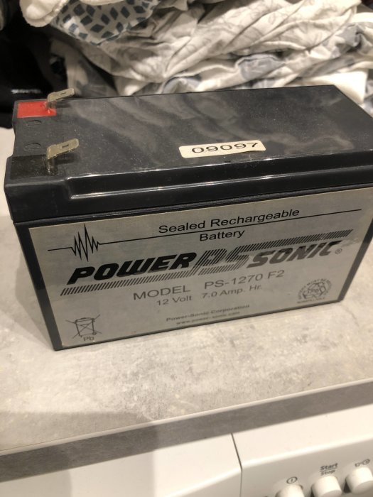 En förseglad laddningsbar Power Sonic blybatteri, 12 volt och 7.0 ampere timmar, står på en arbetsyta.