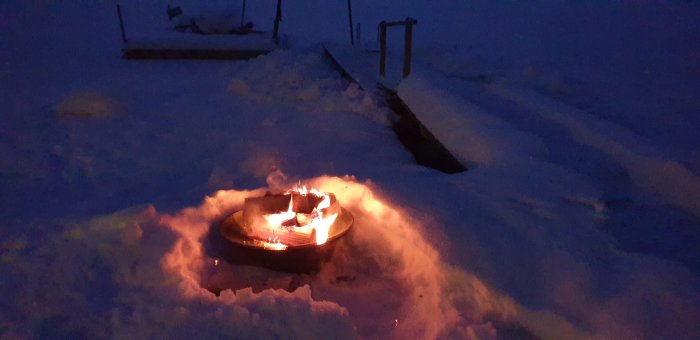 Eldfat med brinnande ved upplyser snön vid en sjökant i skymningen.