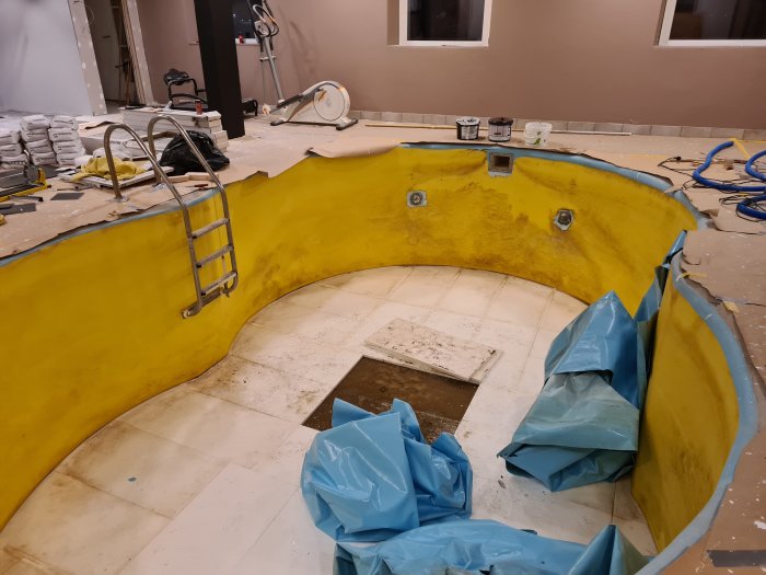 Tömd inomhuspool under renovering, med synlig gul vägg och botten samt borttagen duken, omgivet av renoveringsmaterial.