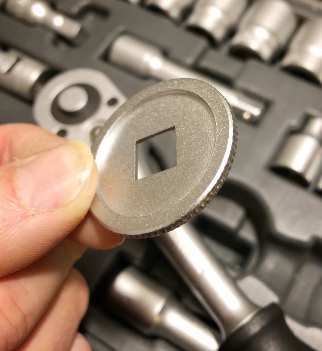 Hand som håller en metallbricka med fyrkantigt hål framför en öppen hylsnyckelsats.