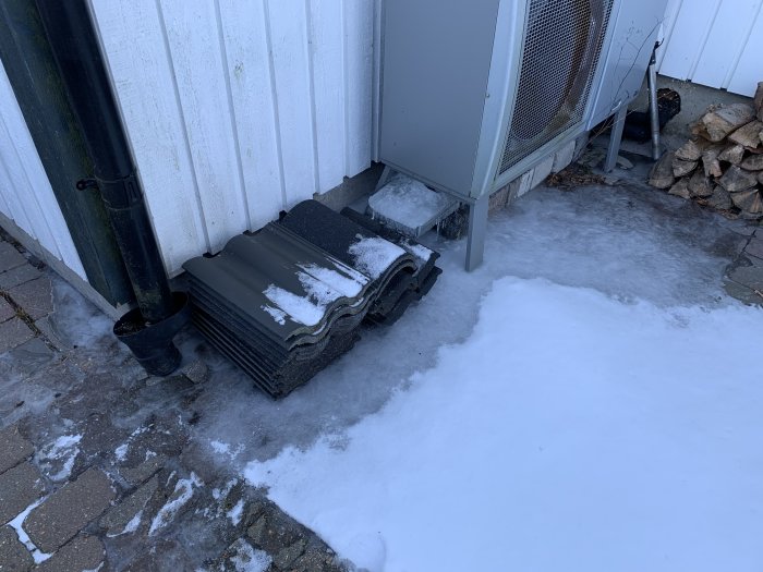 Värmepump med isbildning på marken och avrinningskanal vid husvägg i vintermiljö.