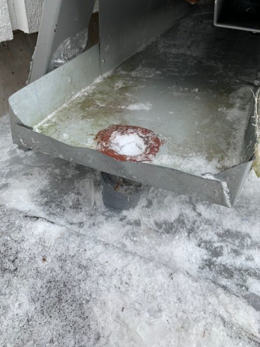 Avtining av isigt tråg med synlig dränering och rostig fläck, placerat över ett fruset plaströr, med snöig yta runtomkring.