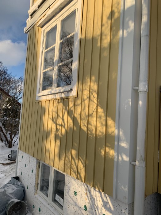 Gult plåtfasad på ett hus från 70-talet med synliga skador nära bottensektionen, omgivet av snö.