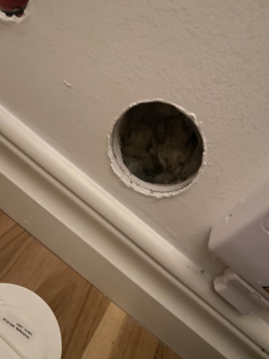 Ett rundat hål i en vit vägg ovanför en golvslist med synlig isolering, nära en doslock och ett eluttag.