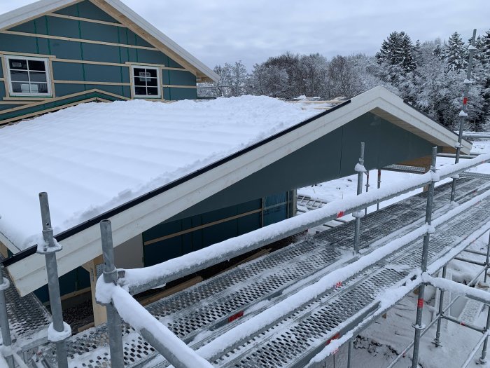 Hus under konstruktion med snötäckt tak, pannbrädor och presenning, synligt reglararbete, i vinterlandskap.