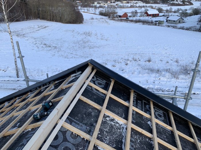 Tak under konstruktion med reglar och takpapp, täckt av lätt snö och bakgrund med snötäckt landskap.