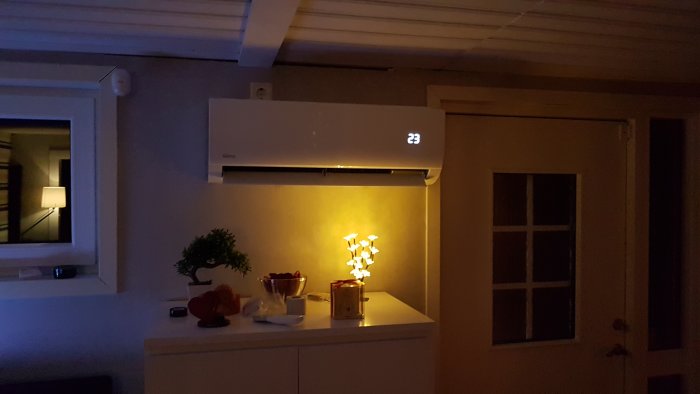 Väggmonterad luftkonditioneringsenhet på 23 grader i ett mörkt rum med dekorativ belysning och fruktskål.