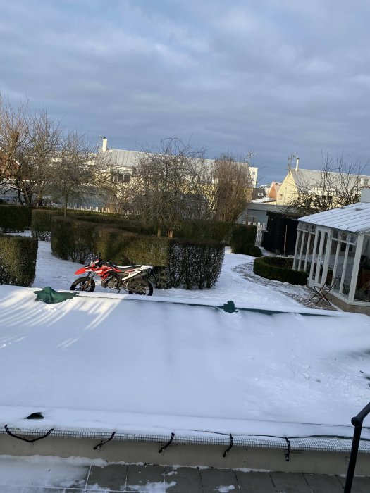 En bakgård med begränsade snömängder och en parkerad crossmotorcykel i dagsljus.