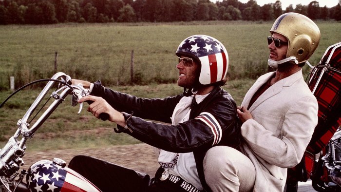 Två män på motorcykel, en med patriotisk hjälm och en i guldhjälm, debatterar hjälmbruk.