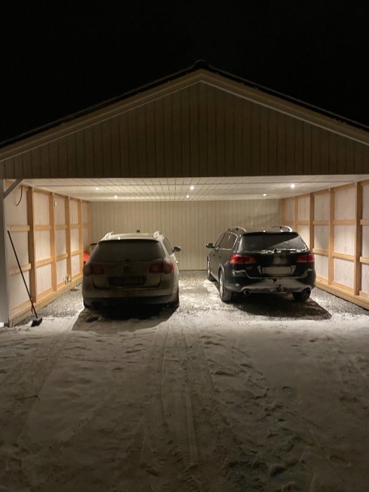 Två bilar parkerade under en carport med synlig takkonstruktion och sidoväggar på natten.