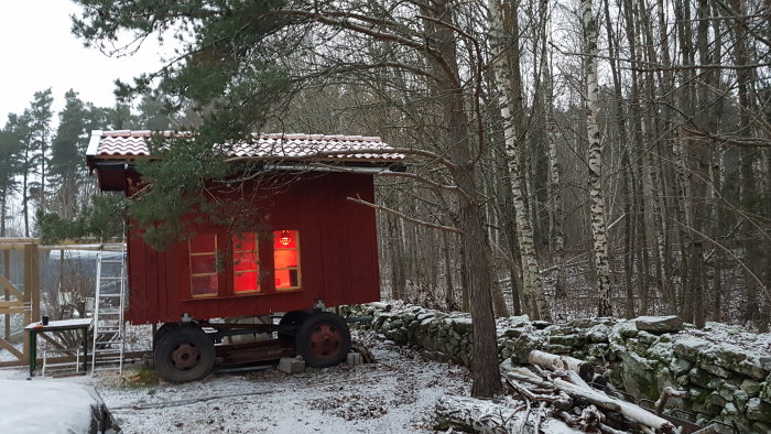 Rödmålat hus på släpvagn med tända fönsterlampor, omgivet av snötäckt skog och stenmur.